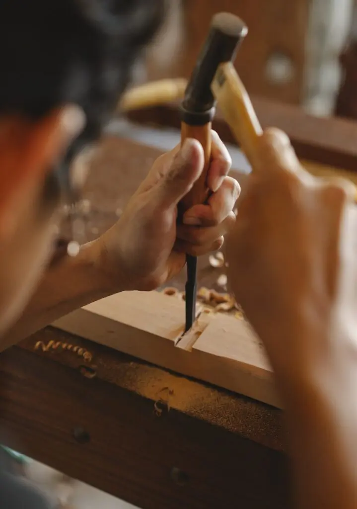 Crop focused artisan making notch in hardwood plank using chisel and small hammer